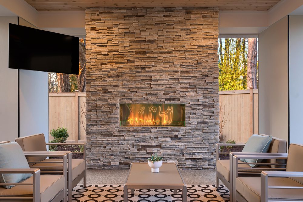 Luxury outdoor relaxing living room with large stone fireplace, TV, rug and beige sofa.