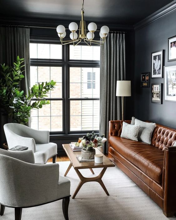 A cozy living room features dark gray walls, a brown leather sofa, two light gray armchairs, and a wooden coffee table. a gold chandelier hangs above, near draped windows and various framed artworks on the walls.