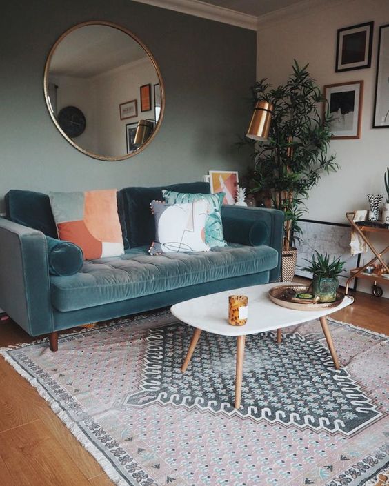 A cozy living room featuring a teal sofa adorned with peach and white cushions, a round white coffee table, patterned rug, hanging plants, and a large round mirror on a grey wall with framed art.