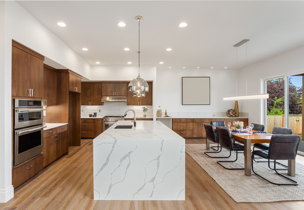 Modern kitchen interior featuring sleek wooden cabinetry, a large white island with marble countertop, built-in stainless steel appliances, and an adjacent dining area with a table set for four. ample natural light fills the room.