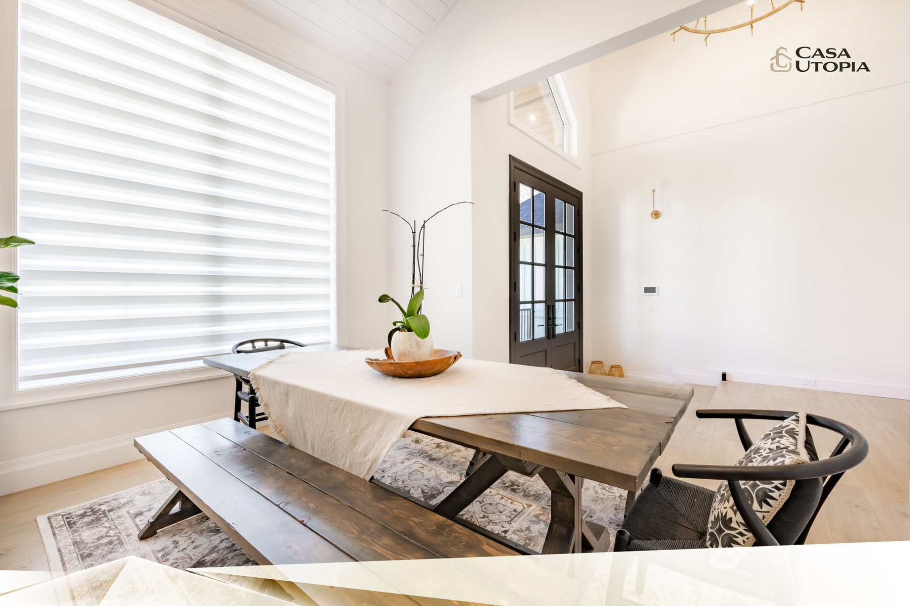 A bright, minimalist dining room featuring a wooden table with benches, patterned cushions, a white orchid centerpiece, and large windows with white blinds. there is a dark door on the right and a wall sconce.