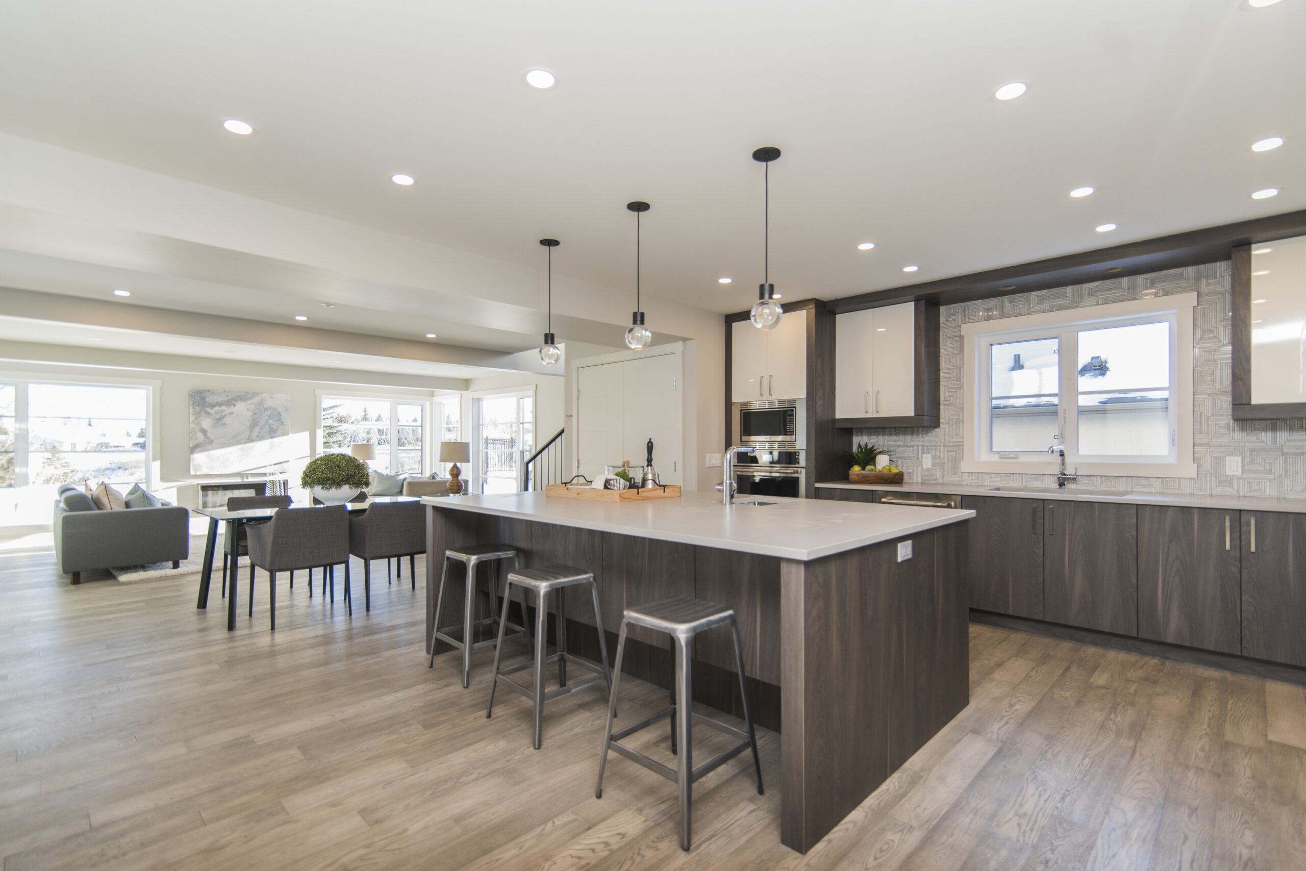 Modern kitchen and dining area with dark wooden cabinets, a marble countertop island with stools, pendant lighting, and stainless steel appliances. the space is bright with natural light from large windows.