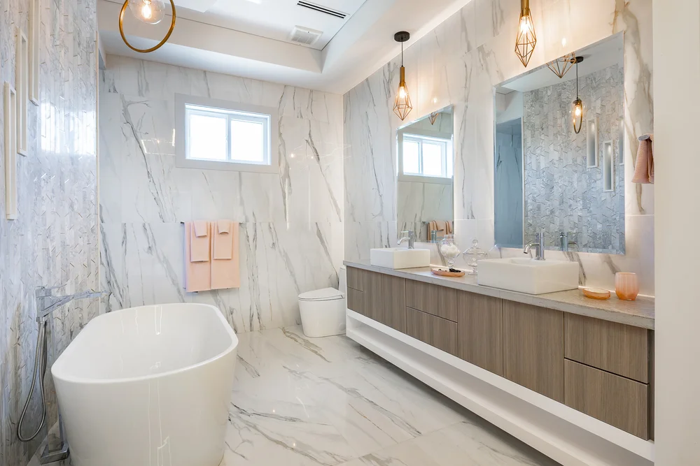 A modern bathroom featuring marble walls and floor, a freestanding white tub on the left, and a long vanity with double sinks. two pendant lights hang beside a large mirror, with coral towels and toiletries arranged neatly. two small windows offer natural light.