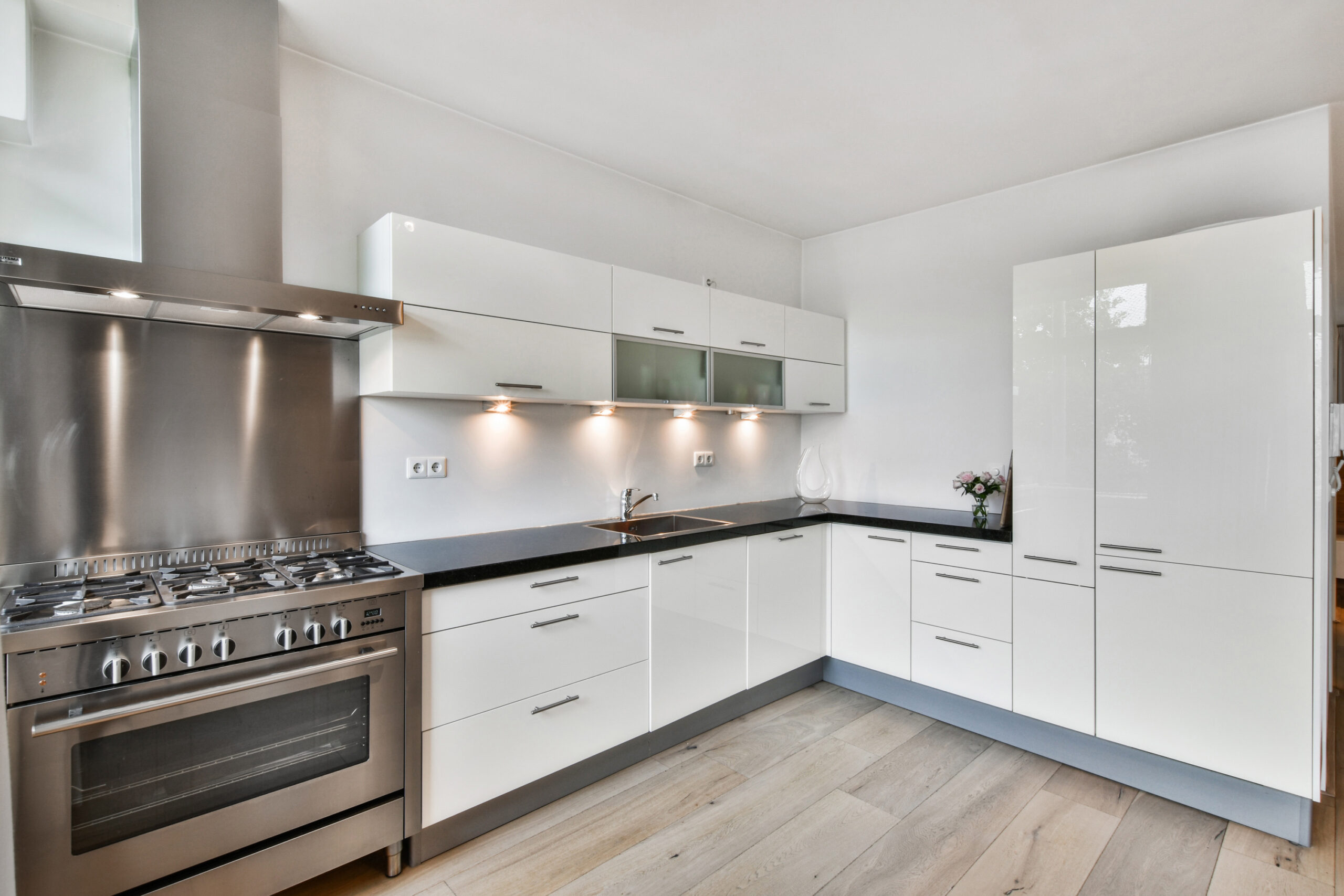 Modern kitchen interior with white cabinetry, stainless steel appliances including a large stove and hood, black countertops, and under-cabinet lighting. light wood flooring complements the clean, sleek design.