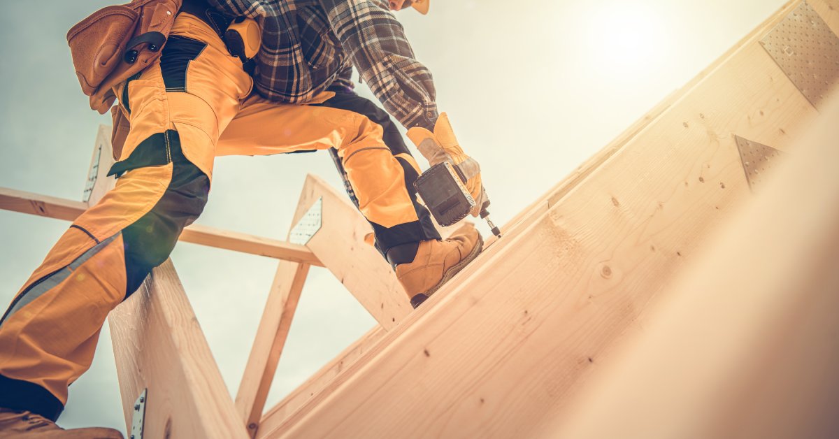 A construction worker wearing safety gloves and a helmet drills into a wooden beam on the ceiling of a home being built.