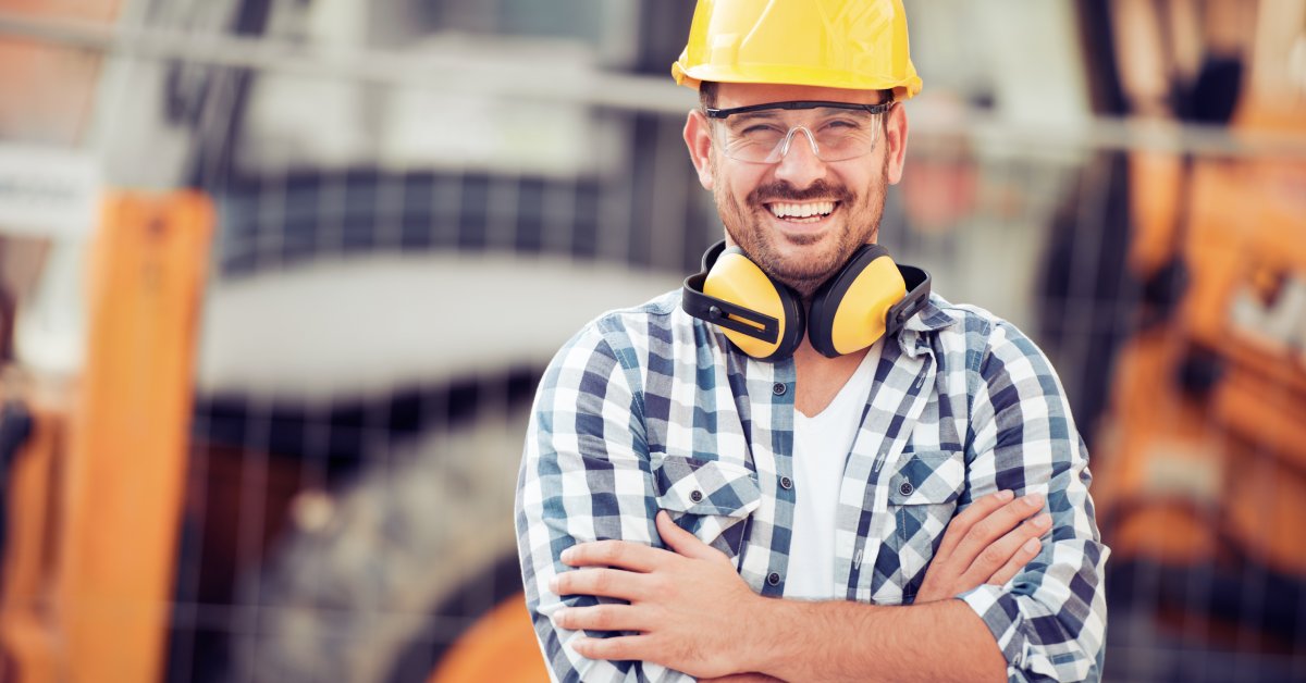 A male contractor wearing a plaid shirt, noise cancelling headphones around his neck, and a yellow helmet.