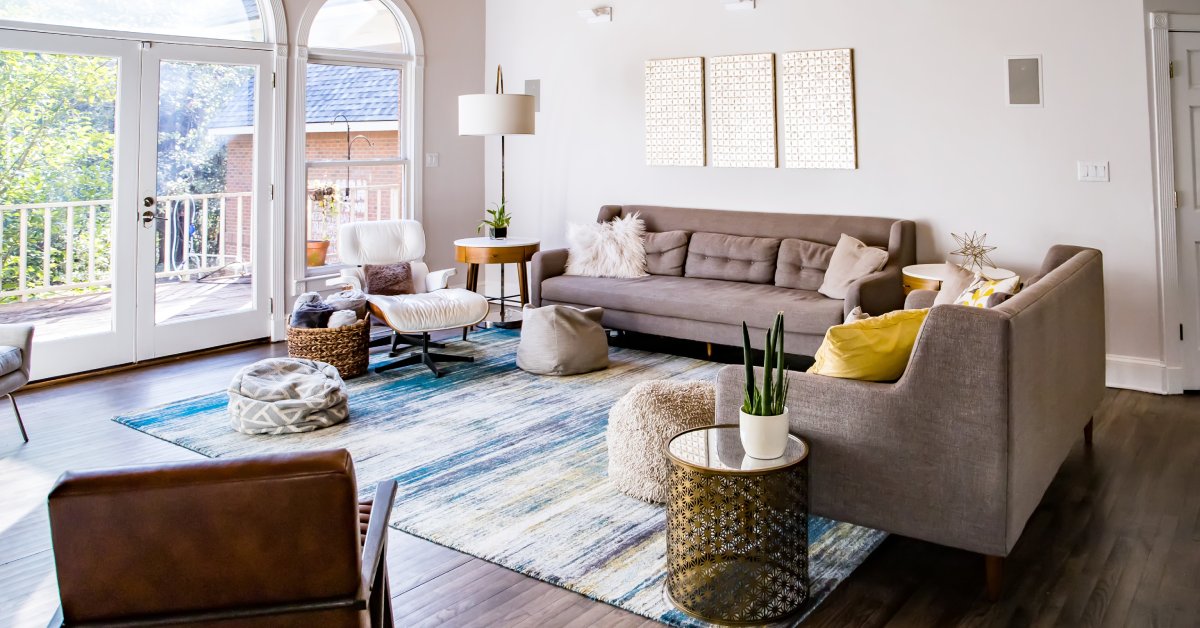 Living room of a mid-century home with wooden floors, brown double couches, a large blue area rug, and large windows.