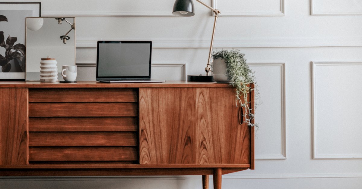 Wooden credenza with sliding doors holding a plant, a lamp, a mirror, a cup, and a laptop placed infront of a white wall.