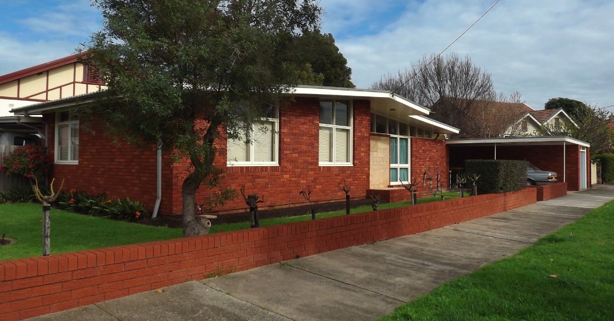 Mid-century one-story home with brick walls, large windows, white frames, a small brick fence, and lush grass.