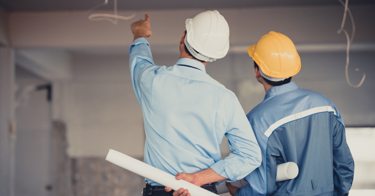 Two men wearing helmets stand inside a room under construction, while looking at the ceiling and holding large rolls of paper.