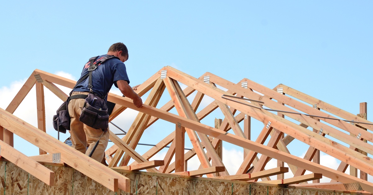 A construction worker wearing a tool belt and a blue t-shirt standing on top of a large wooden home structure.