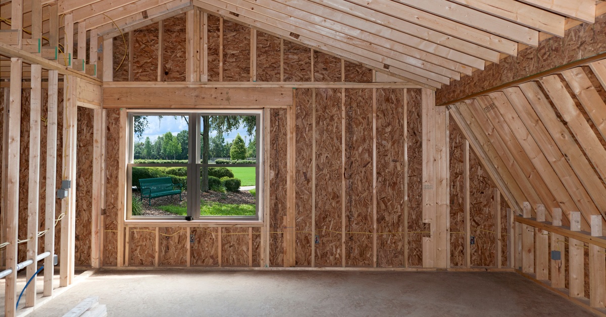 A room with wooden boards, exposed wooden beams and electrical wiring, with a small window overlooking grass.