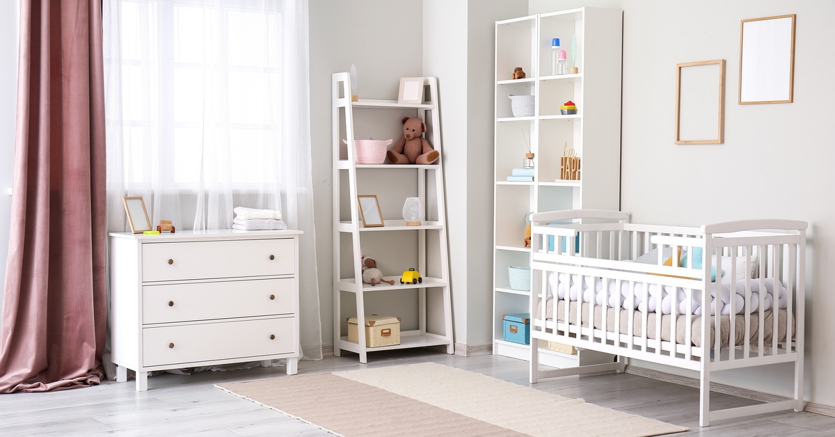 A room with white walls, a crib, a small dresser, a rug, stuffed animals, two bookshelves, and a small window.