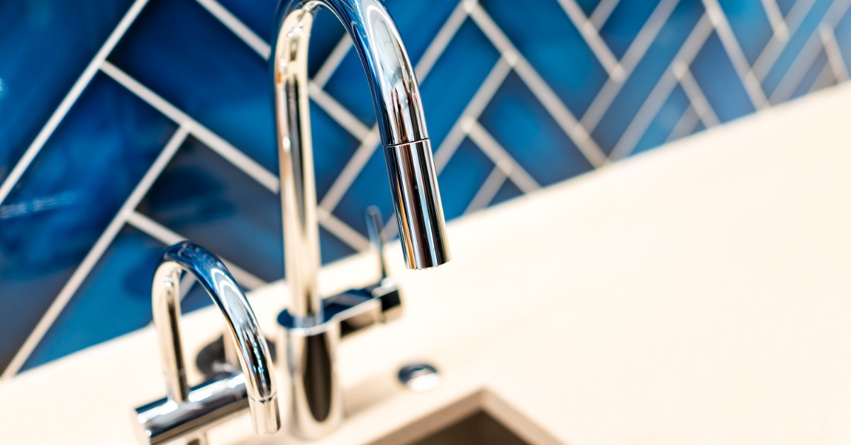 A close-up of a curved stainless-steel faucet next to a small faucet on a sink with bright blue tiles for a backsplash.