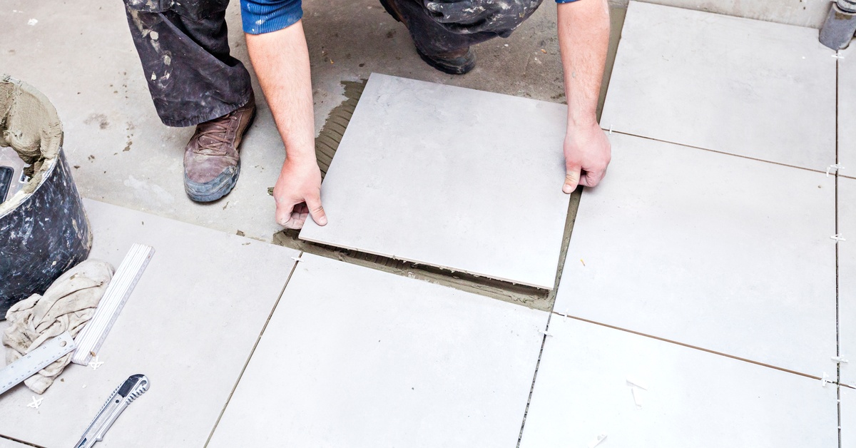 A close-up of the hands of a man placing a large, white square tile over wet cement on the floor next to other tiles.