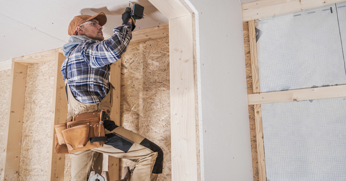 A man wearing a tool belt and safety glasses, standing on top of a folding ladder, drilling a hole in a ceiling panel.