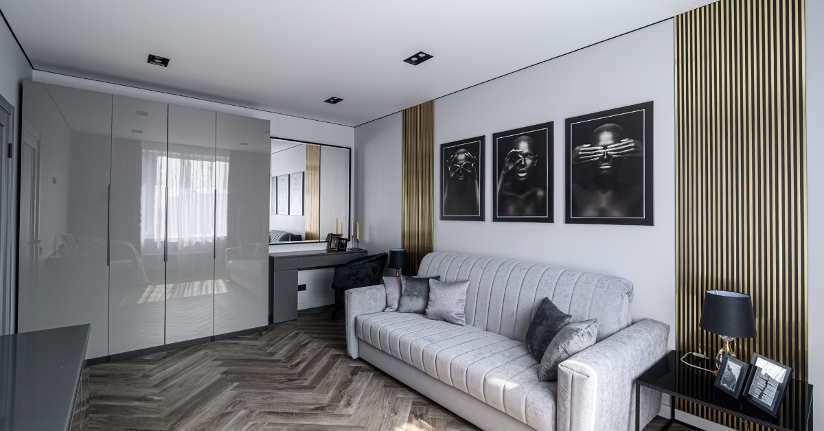A room with wooden floors, wooden gold panels on a white wall, a grey couch, a small desk, and a closet.