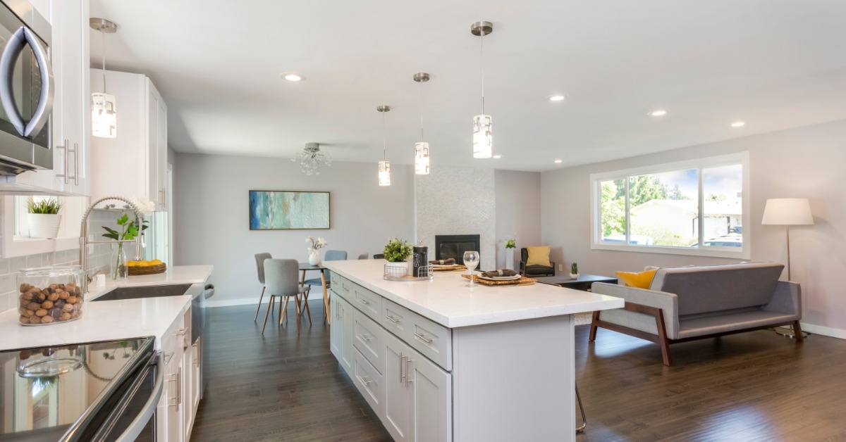 A kitchen with a large white island, next to a living room with a brown couch, a large window and a dining table.