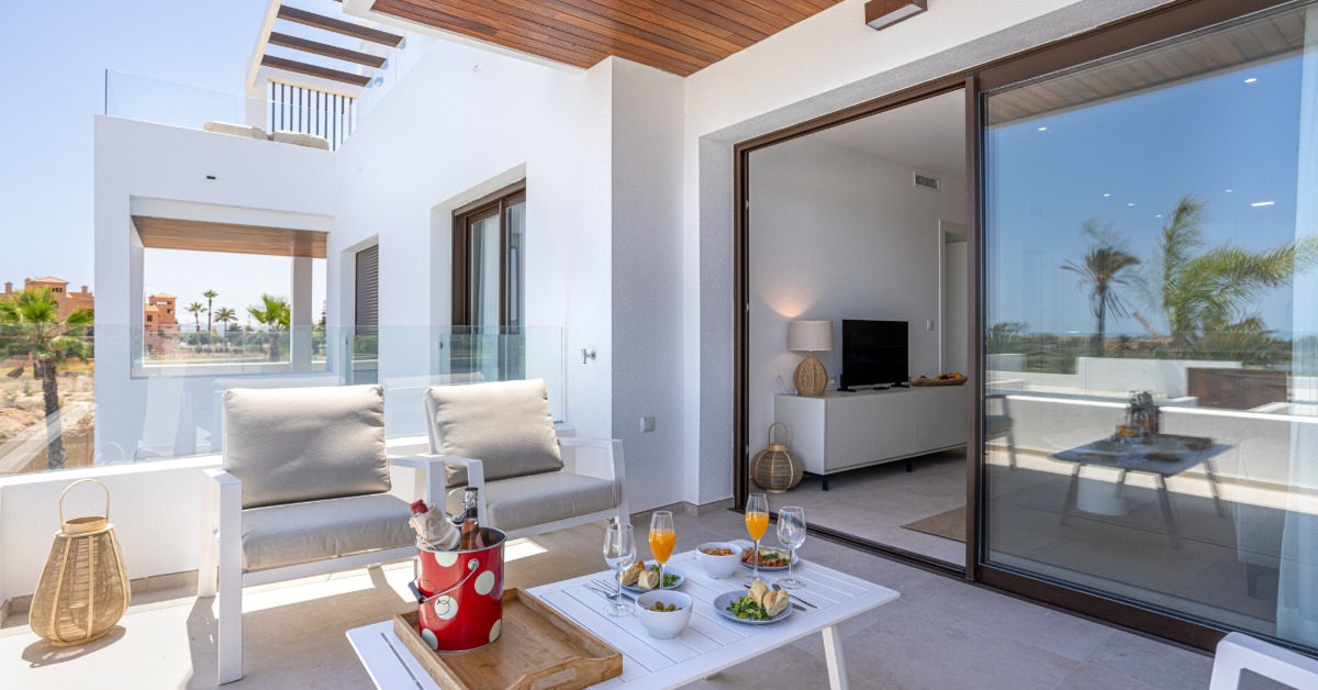 An outdoor living space with chairs and a table, with an open sliding glass door connecting to an interior living room.