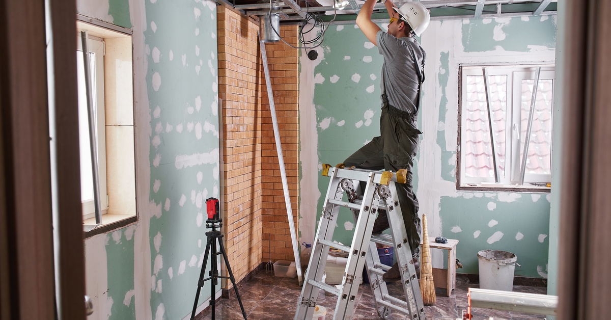 A man wearing a white helmet, green overalls, and gloves, standing on a ladder inside a room, touching a metal beam.