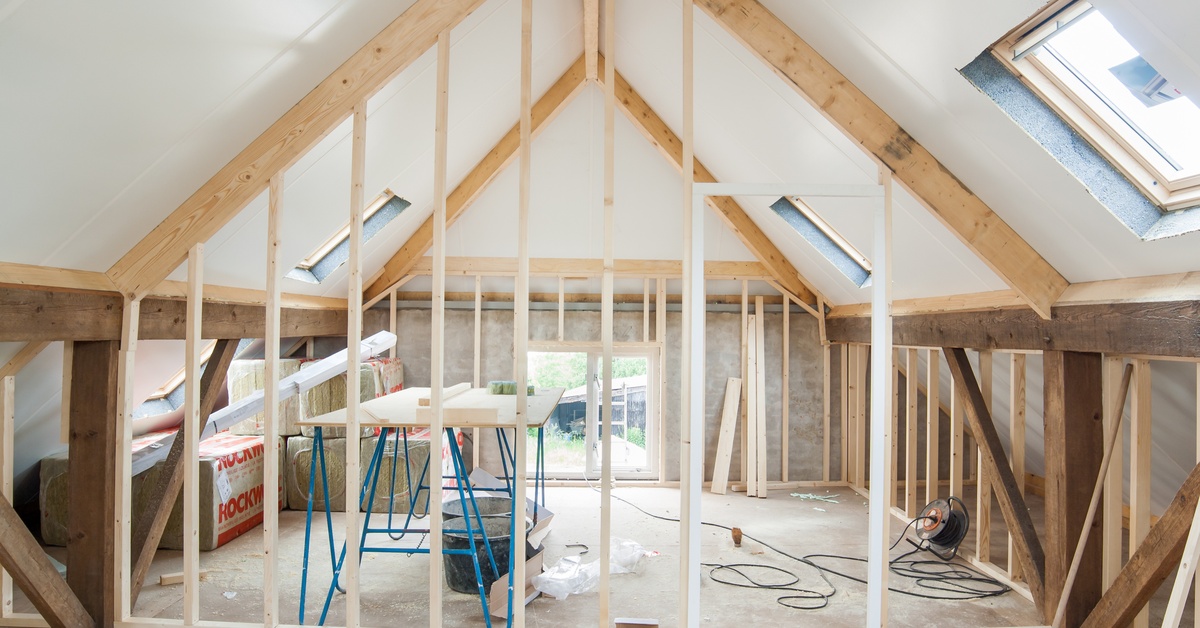 A room undergoing renovations with a sharp roof, three skylights, multiple wooden beams, a table, and brooms.