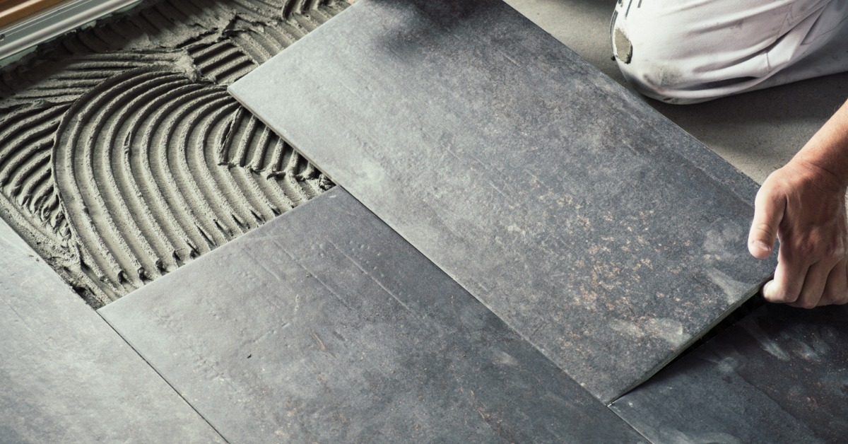 A close-up of the hands of a worker laying a large rectangular black tile on a floor covered with cement adhesive.