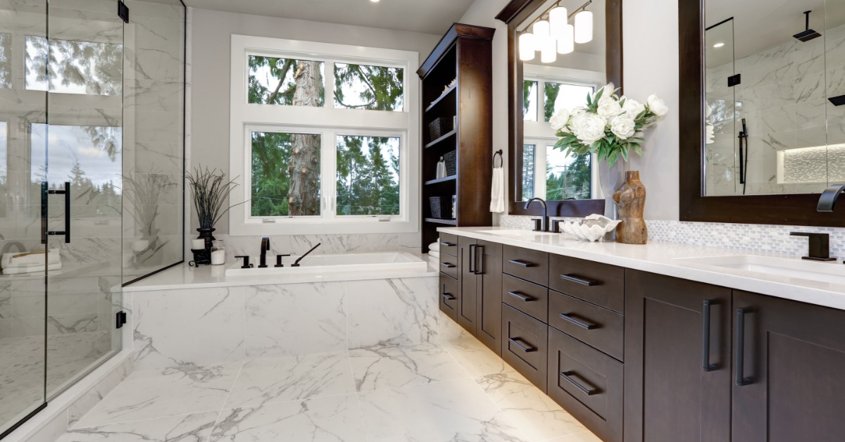 A luxurious bathroom with marble floors, brown cabinets, double sinks, a walk-in shower, and a tub with a bookshelf.