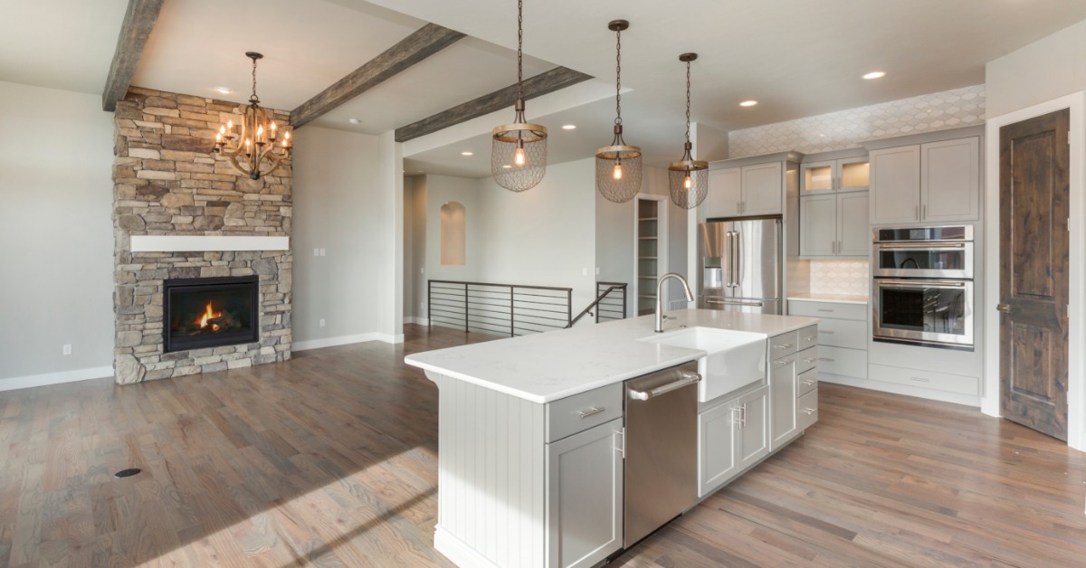 A kitchen with wooden floors, a white island, grey cabinets, and stainless-steel appliances next to the living room.