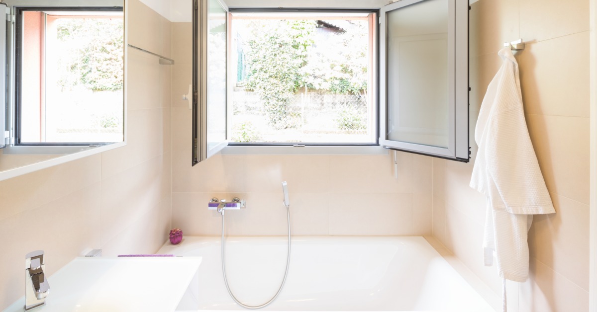 A bathroom with light yellow walls, a white sink, a mirror, and a white tub in the back under a large open window.