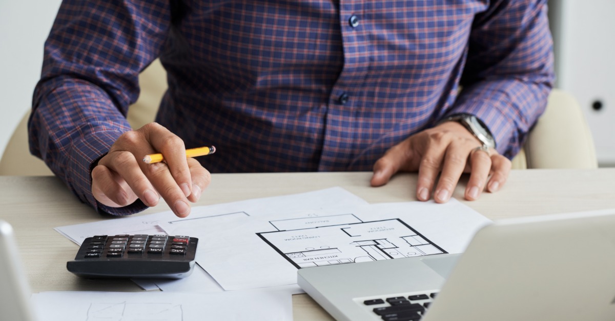 A man sitting behind a white desk with construction plans, holding a pencil, using a calculator and a laptop.