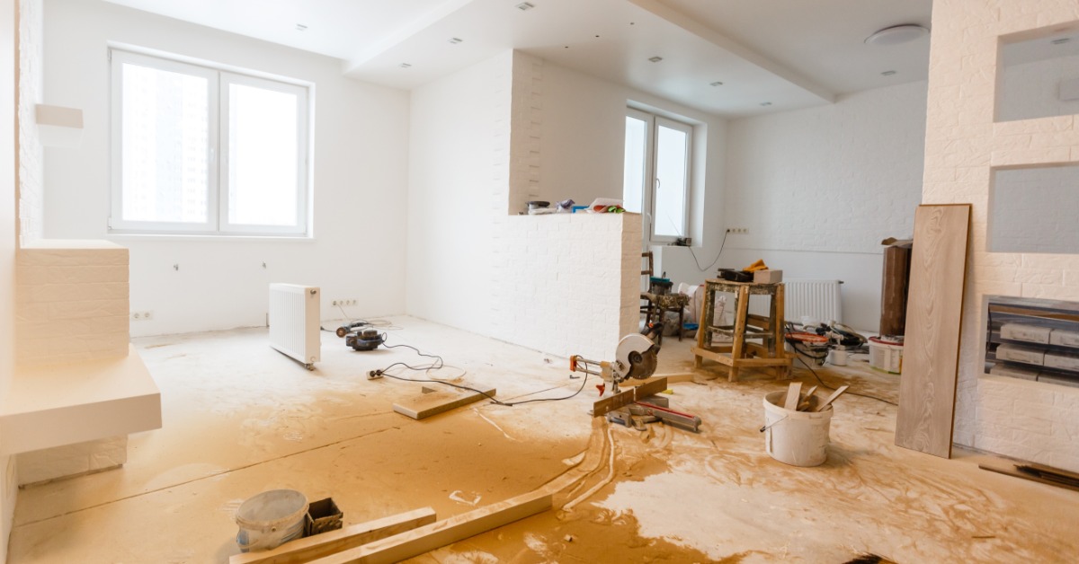 A large room undergoing a renovation with multiple tools, wooden beams, a broom, and wood dust on the floor.