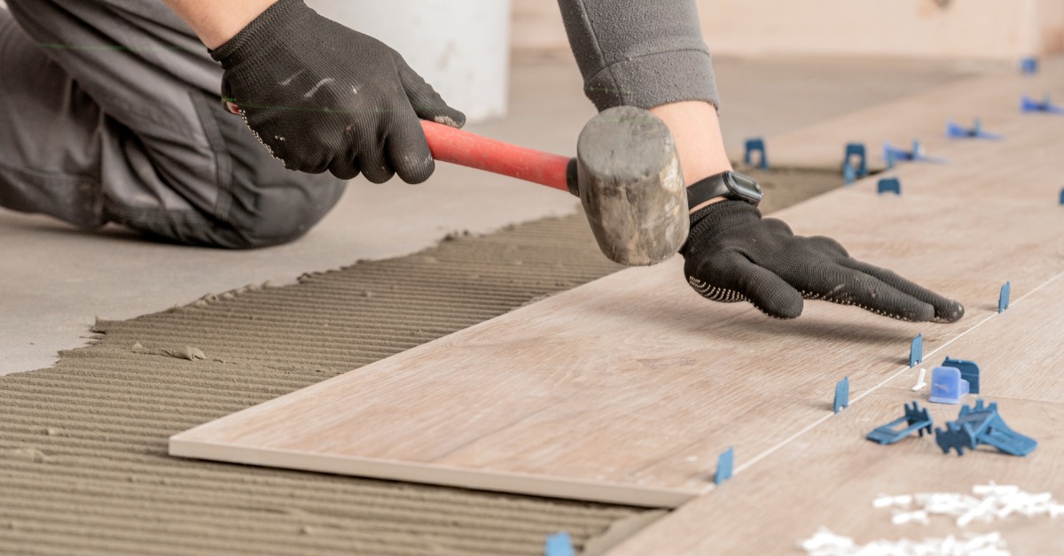 A close-up of two hands wearing black protection gloves, holding a hammer while touching a beige tile on the floor.