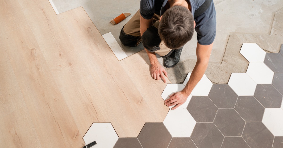 An aerial view of a man kneeling on the floor while placing a hexagon tile next to other tiles and laminated strips.