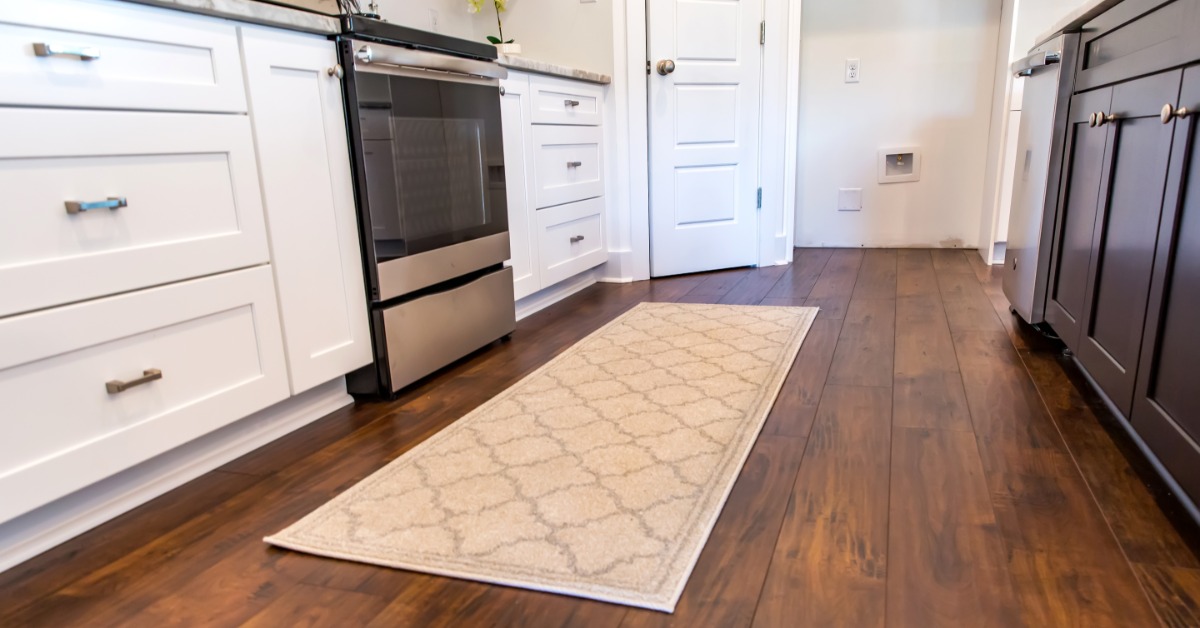 A close-up of a wooden floor with a strip rug inside a kitchen with stainless steel appliances and white cabinets.