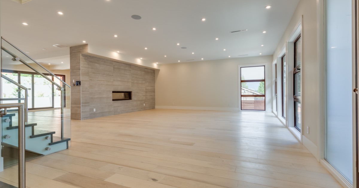 A large empty room with wooden floors, white walls, multiple overhead lights, and floor-to-ceiling windows.