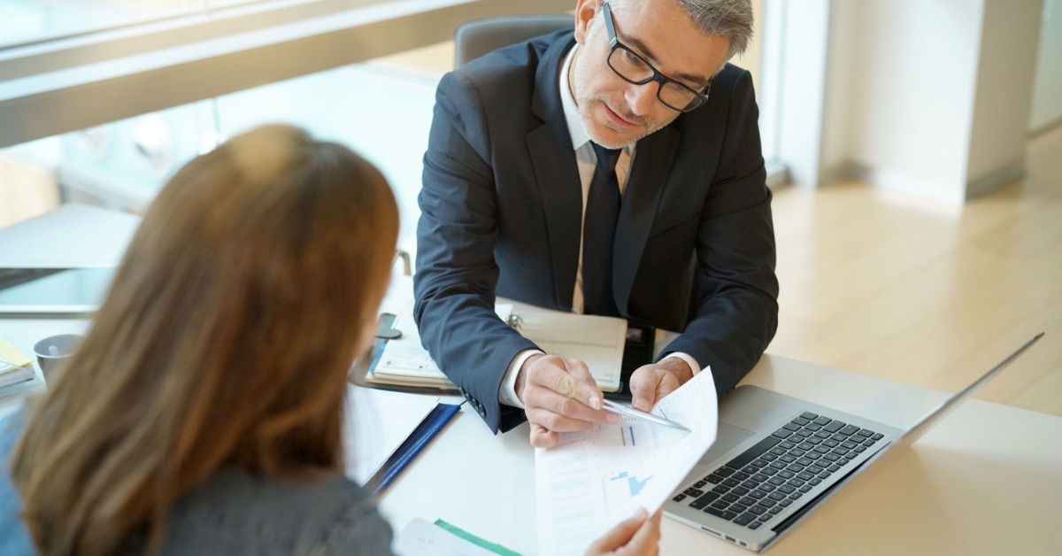 A man sitting behind a desk holding a sheet of paper and pointing to a specific location to a woman in front of him.