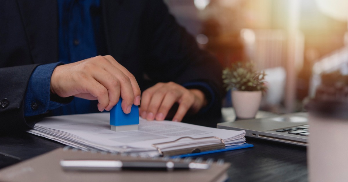 A close-up of a man stamping a document placed on a clipboard resting on top of a black desk next to a laptop.