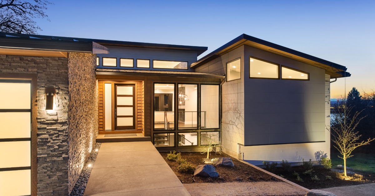A one-story home with a concrete entrance pathway, rock walls, a small front garden, and ample lighting during a sunset.