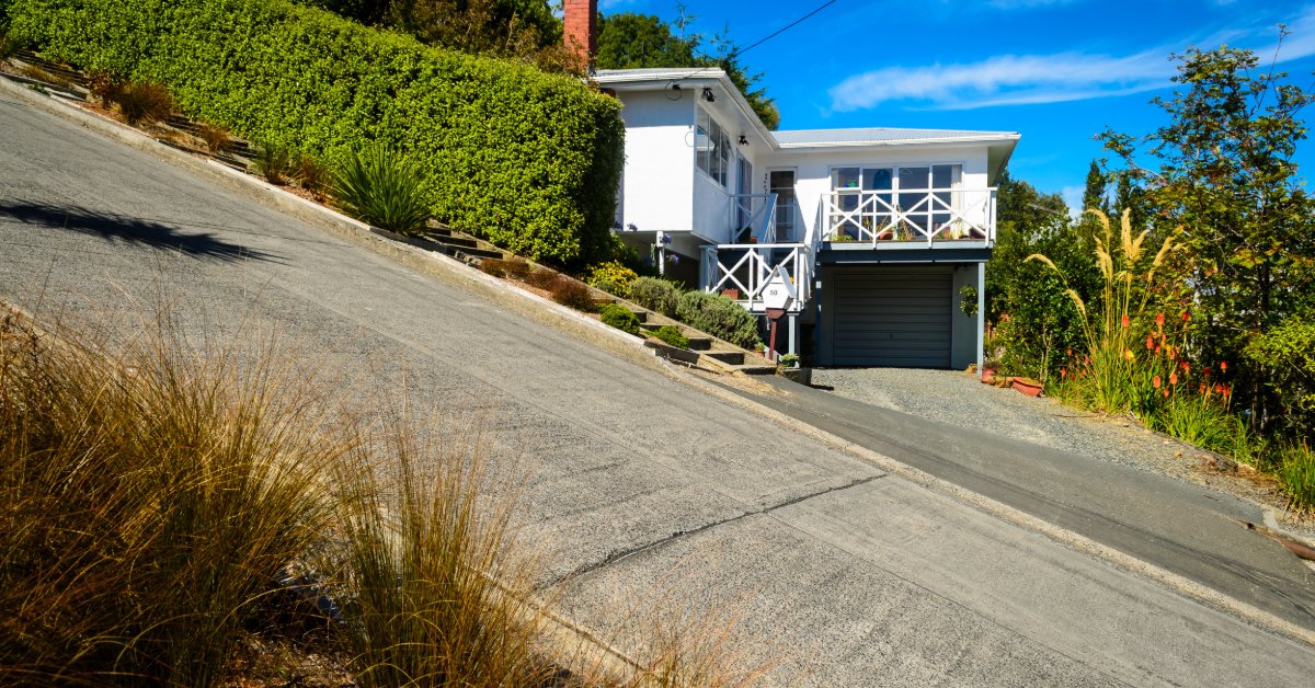 A two-store white house with an exterior staircase, a chimney, and trees, built on a steep concrete street.
