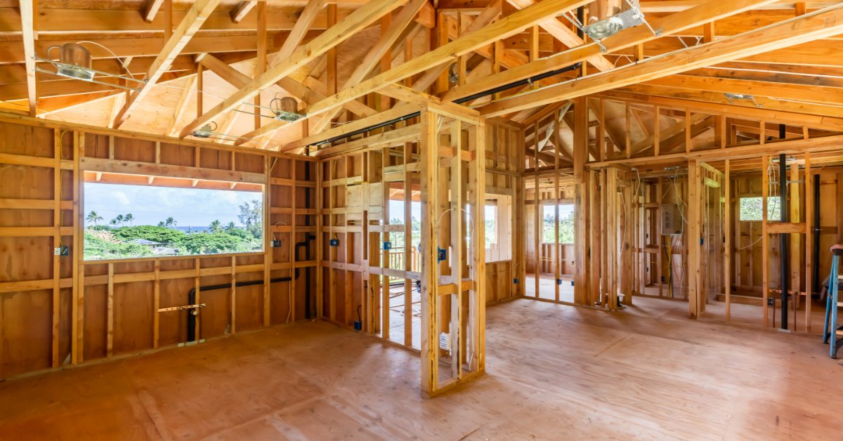 A large room with exposed wooden beams on the walls and ceiling, with multiple windows overlooking greenery.