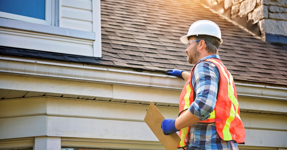 A worker wearing a safety vest, a helmet, and latex gloves, touching a shingle on the edge of a house roof.