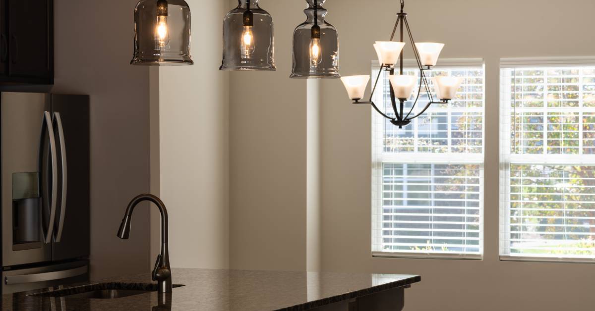 A metal chandelier with seven lightbulbs hanging next to another chandelier with three glass cases in a kitchen.
