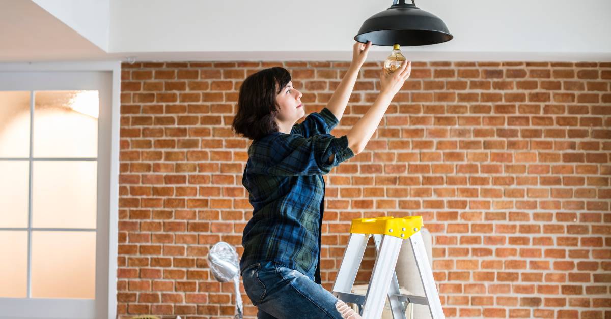 A close-up of two hands wearing safety white gloves, installing a long white lightbulb in the ceiling of a room.