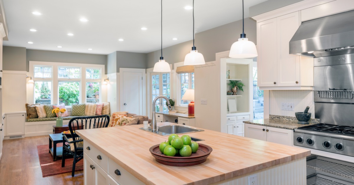A kitchen with a large island, stainless steel appliances, and three hanging lights, next to a colorful living room.