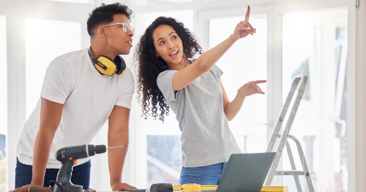 A woman pointing at the ceiling of a room under renovation, next to a man leaning on a table with construction tools.