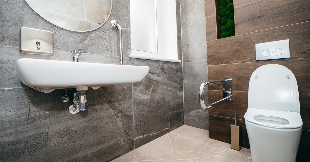 A bathroom with brown walls, a floating sink, a handrail on the wall next to a toilet, and a small window on the side.