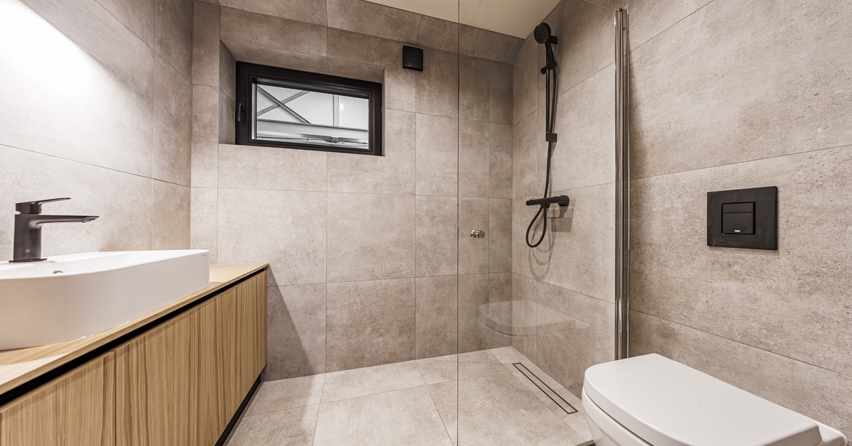 A modern bathroom with a large walk-in shower with black fixtures, next to a white toilet and a long sink counter.