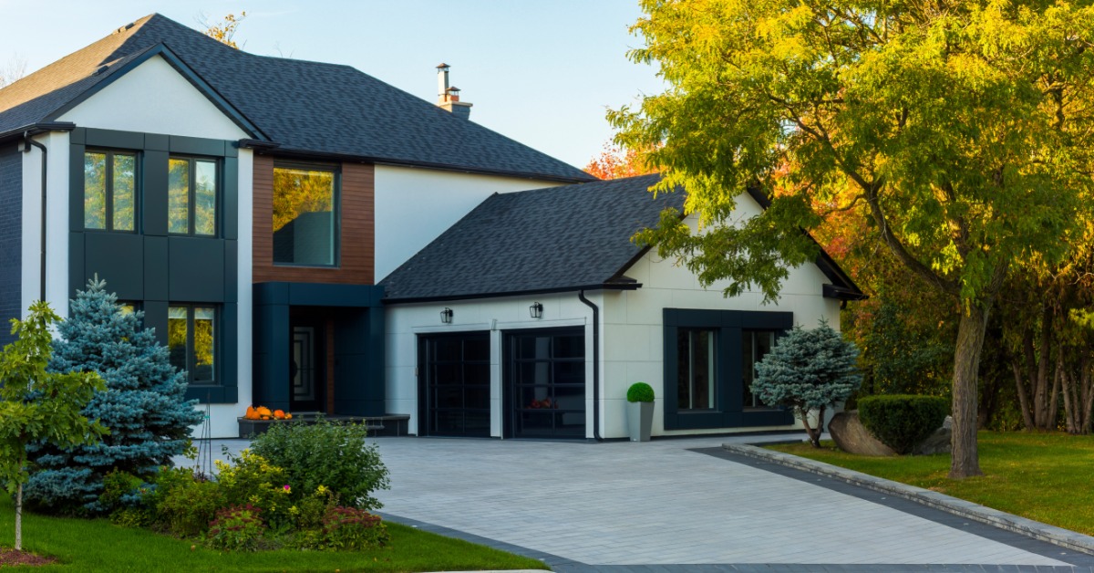 A two-story home with a double garage, white walls, and a roof with grey shingles, surrounded by greenery.