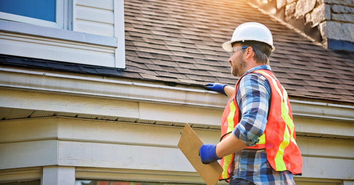 A man wearing an orange vest and a white helmet touching the side of a residential roof with asphalt shingles.