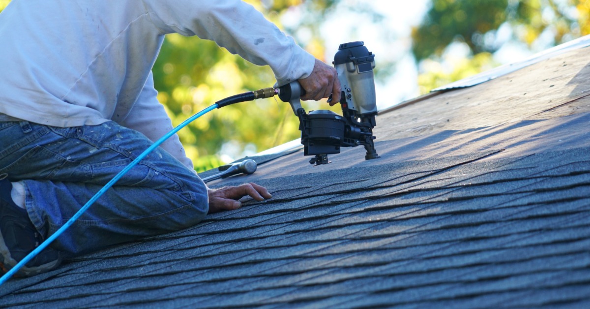 A man wearing a white shirt, kneeling on top of a residential roof, using a pneumatic nail gun to attach shingles.
